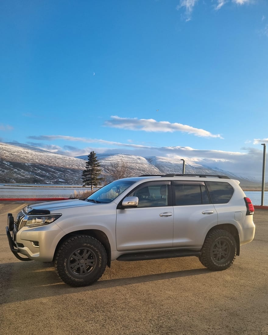 Silfurlitaður jeppi Jóns undir bláum himni með snævi þakin fjöll í bakgrunni